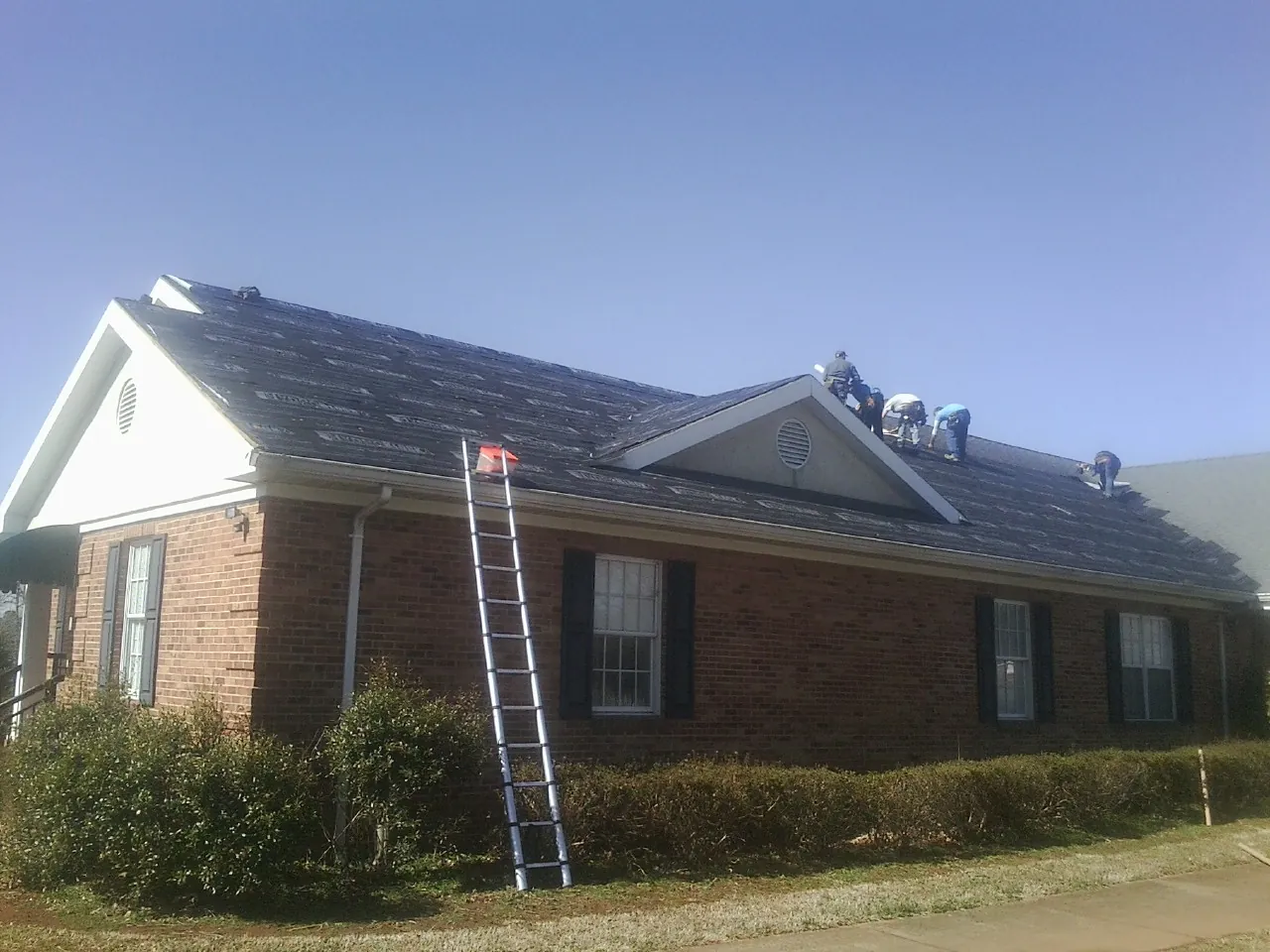 Crew tearing shingles off a brick ranch roof with a ladder set — Metal Roof Repair work by our crew in Dover AFB